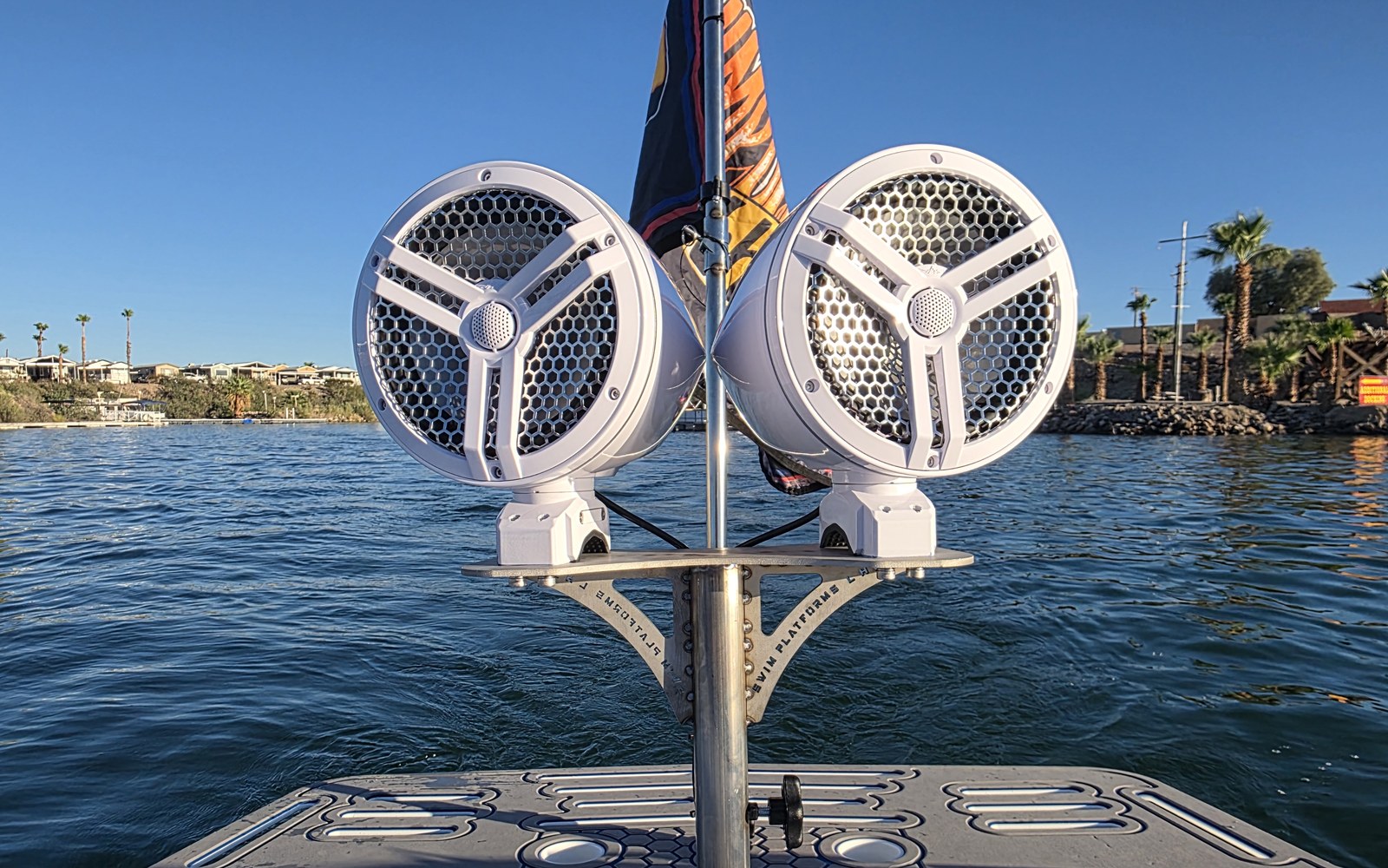 Pair of white pod-style marine speakers on a brushed stainless steel bracket mounted on a post above gray marine decking, with open lake water behind