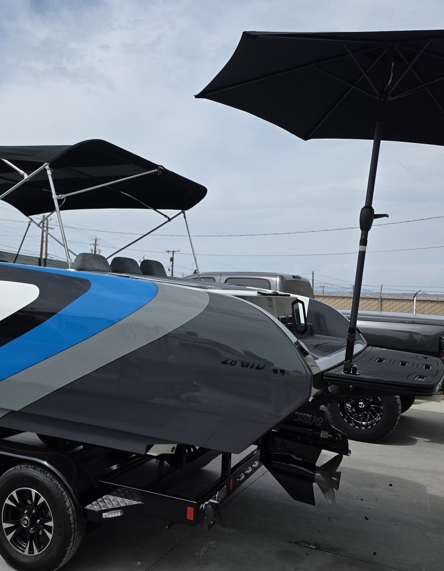 Black and silver performance boat on a trailer outside the shop, seen from the stern, with a black Sandbar Shade umbrella extended over its swim platform
