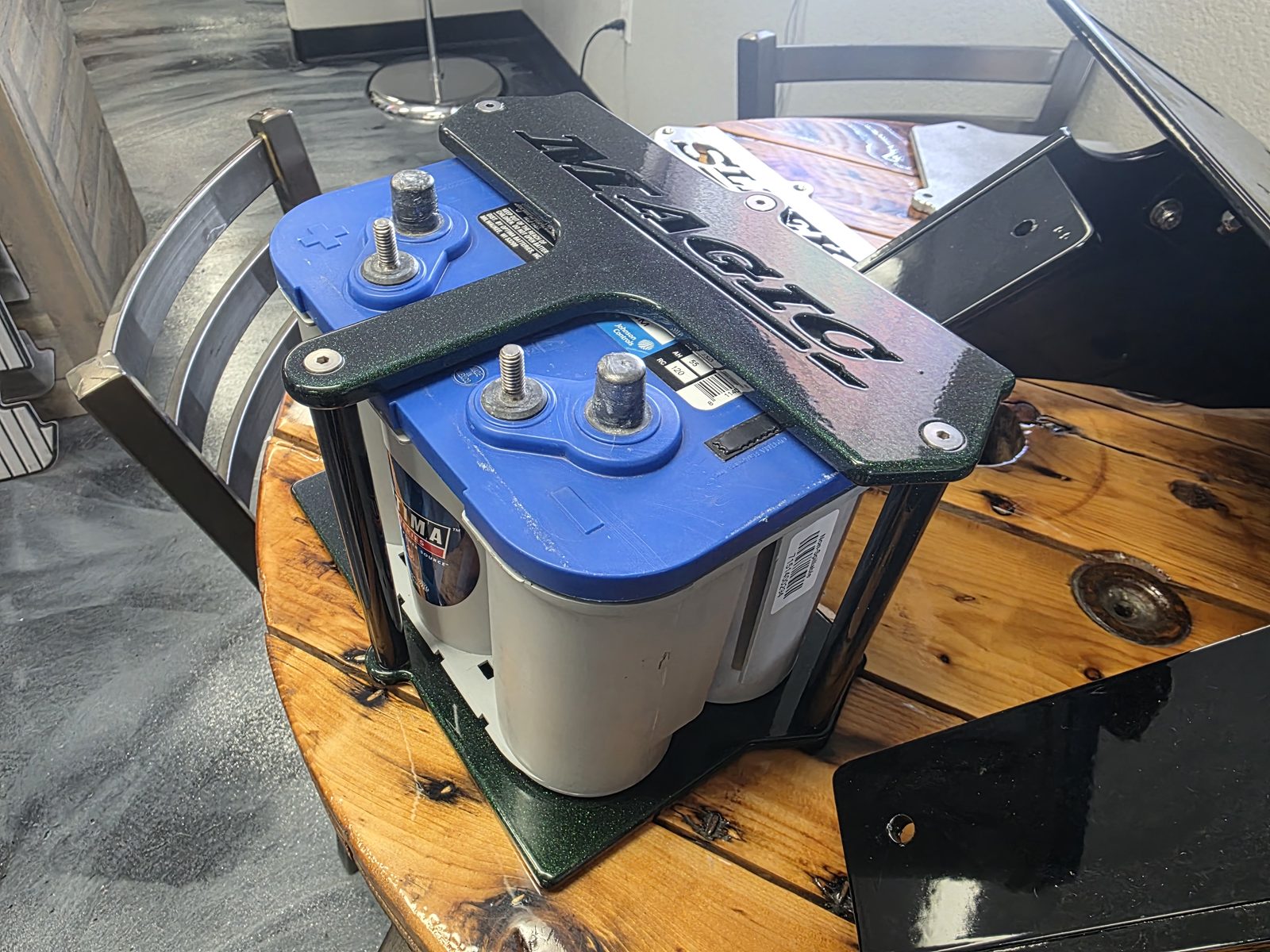 Optima Blue Top battery held in a custom black powder coated aluminum tray with MAGIC cut into the top brace, sitting on a wooden shop table