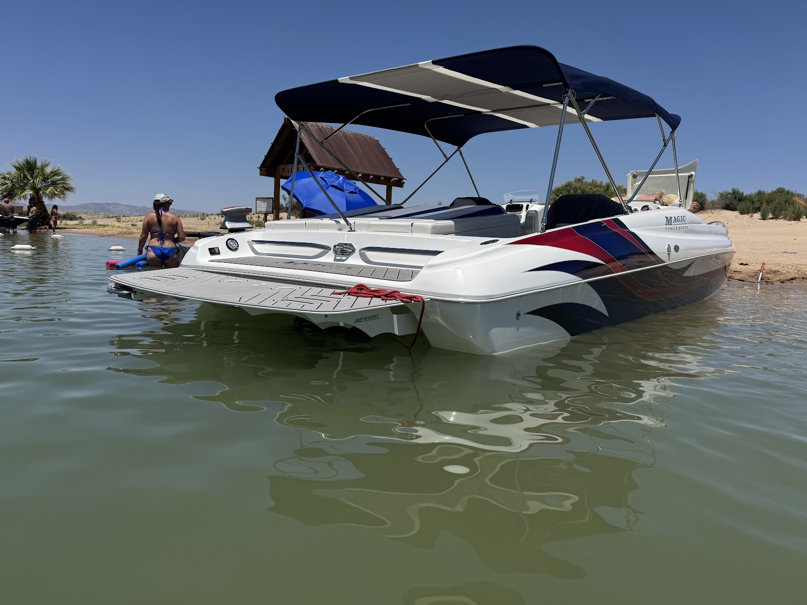 White deck boat with a black bimini top anchored in clear shallow water